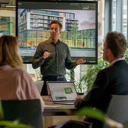 Man presenteert architectuurvisualisatie op groot scherm aan twee collega’s met laptops. Hij legt enthousiast het ontwerp toe in een lichte vergaderruimte.